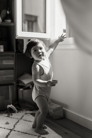 A small child in a leotard reaches up toward a cabinet while standing on a patterned rug in black and white lighting.