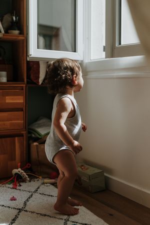 A toddler in a white bodysuit stands near a window in a cozy room with hardwood floors and a patterned rug.