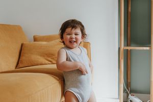 A toddler in a white onesie smiles brightly while standing next to a tan couch against a white wall.