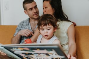 Family sitting together on orange couch looking at photo album with toddler pointing at pages.