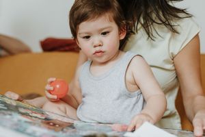 Small child in gray tank top sitting and holding a red toy while looking at a book.