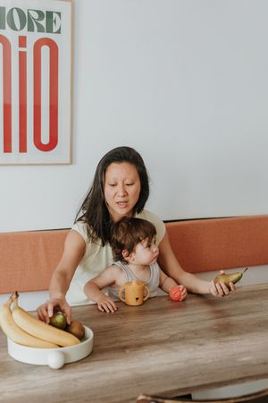 A parent and child sitting at a wooden table with fruits and play toys for a meal or activity.