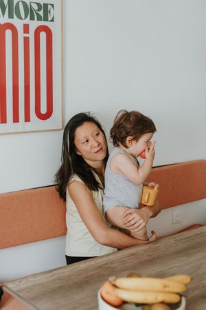 A parent and toddler share a meal together at a wooden dining table with a retro-style sign visible on the wall.