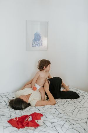 A diaper-clad toddler on bed with patterned linens while someone reclines wearing black and white, with wall art above.