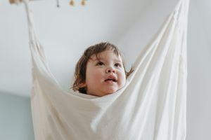 A small child peeks out playfully from white draped fabric hanging from above while looking upward with joy and wonder.
