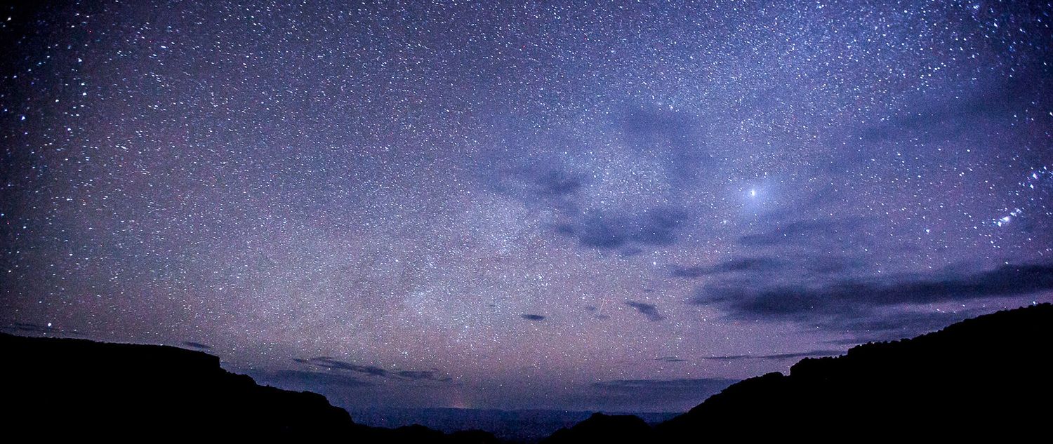 Starry night sky with purple and blue hues over dark silhouetted hills in a panoramic view.