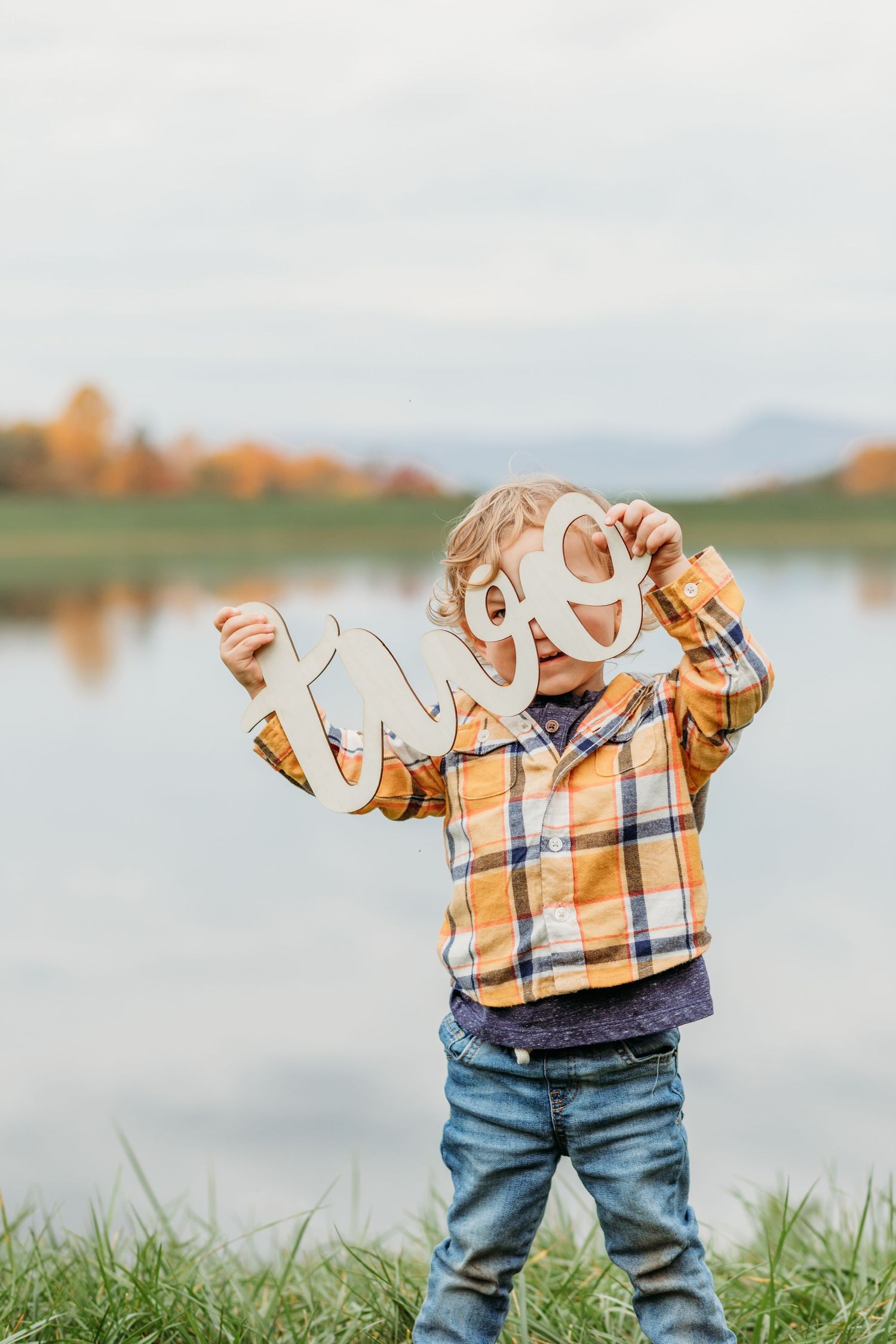 Small child wearing a plaid shirt and jeans plays outdoors by a lake during autumn.