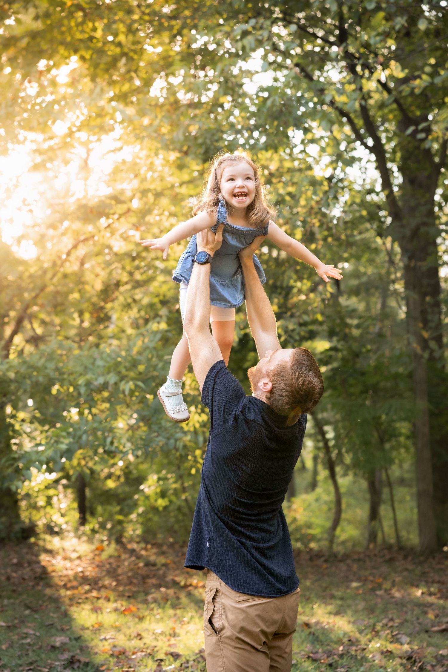 Father joyfully tosses laughing little girl in the air outdoors in a sunlit forest park on a warm sunny day.