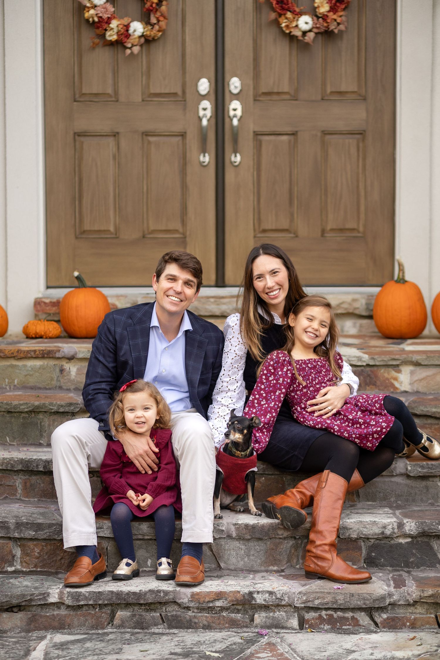 Family of four with small dog posing on fall-decorated front steps with pumpkins and autumn wreaths on wooden doors.
