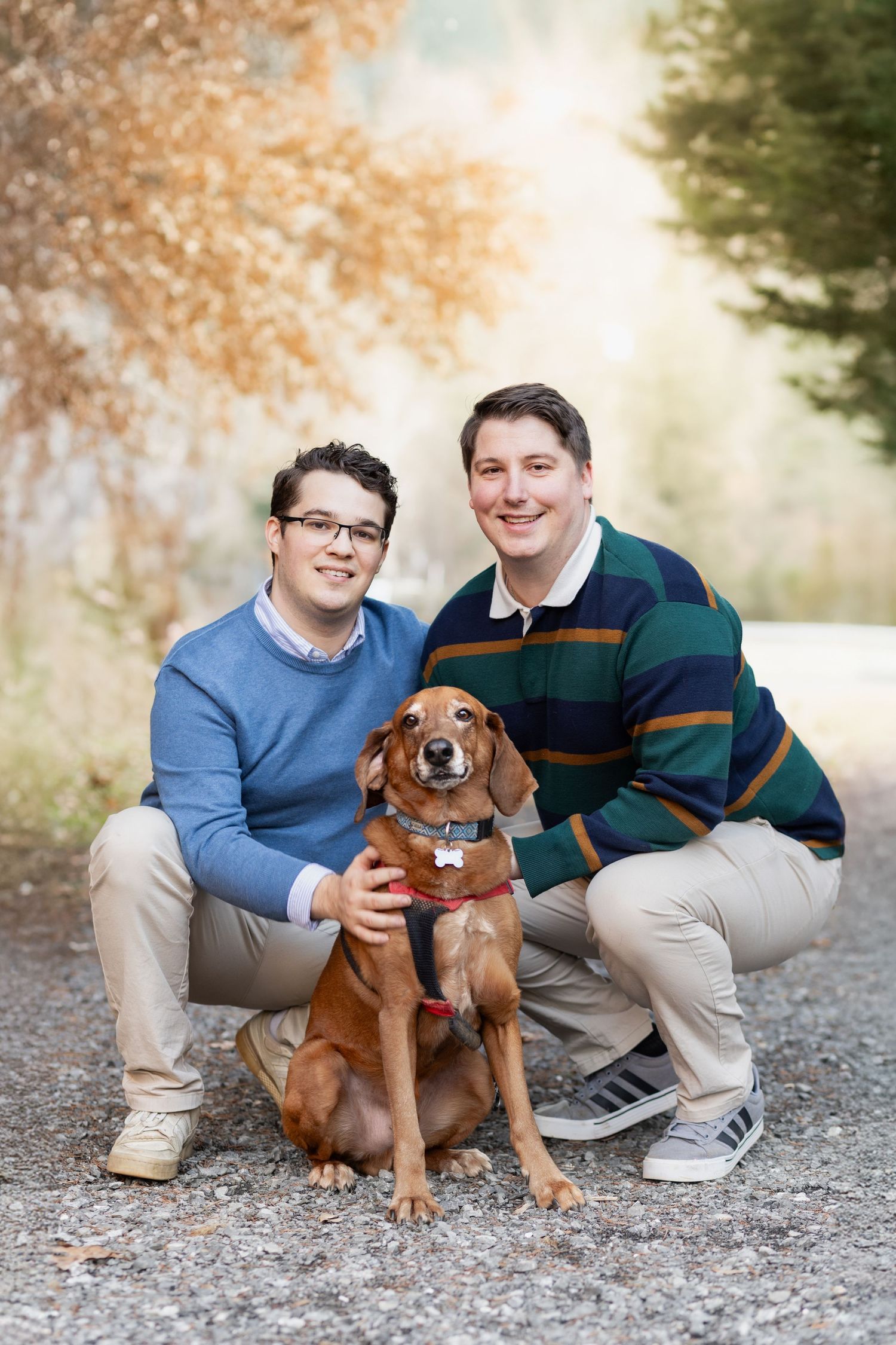 Two smiling men crouch with a brown dog on a gravel path surrounded by autumn foliage.