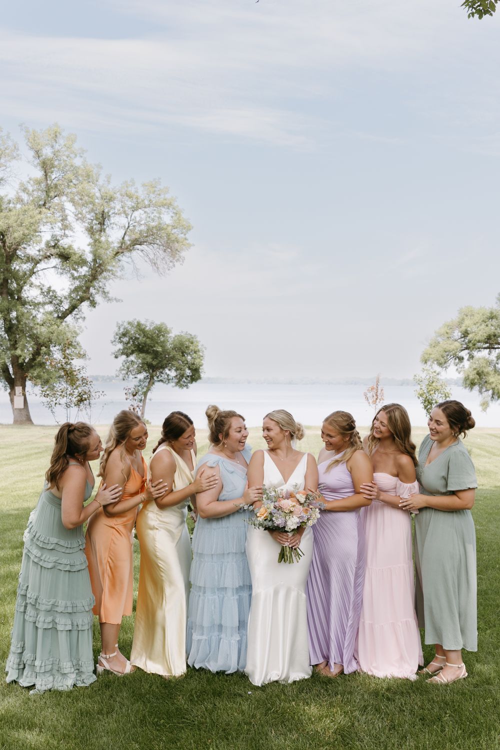 Bridesmaids in pastel dresses gather with bride on grassy lawn.