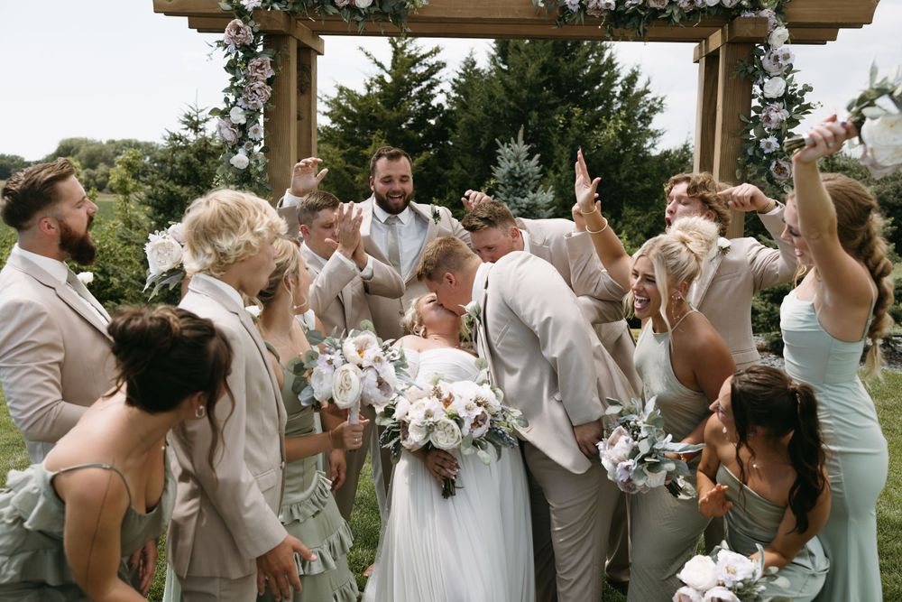 Wedding party cheers and celebrates around the newlyweds under a wooden pergola.