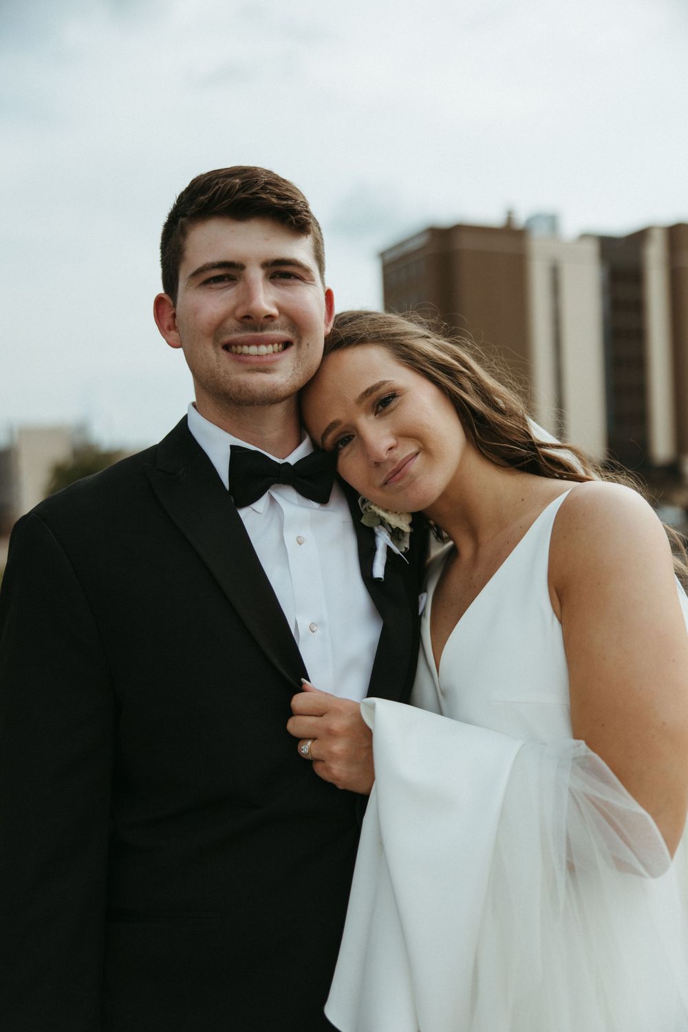 A bride and groom share an intimate moment in formal wedding attire against a city backdrop.