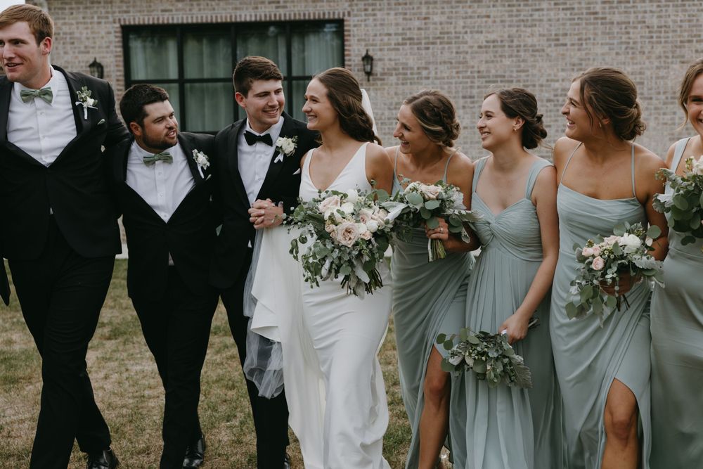 A wedding party walks together outdoors wearing blue bridesmaid dresses and black tuxedos.