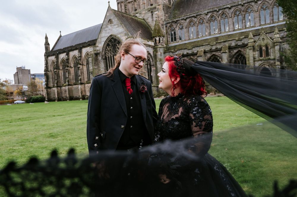Couple poses on a grassy lawn in front of historic cathedral architecture.
