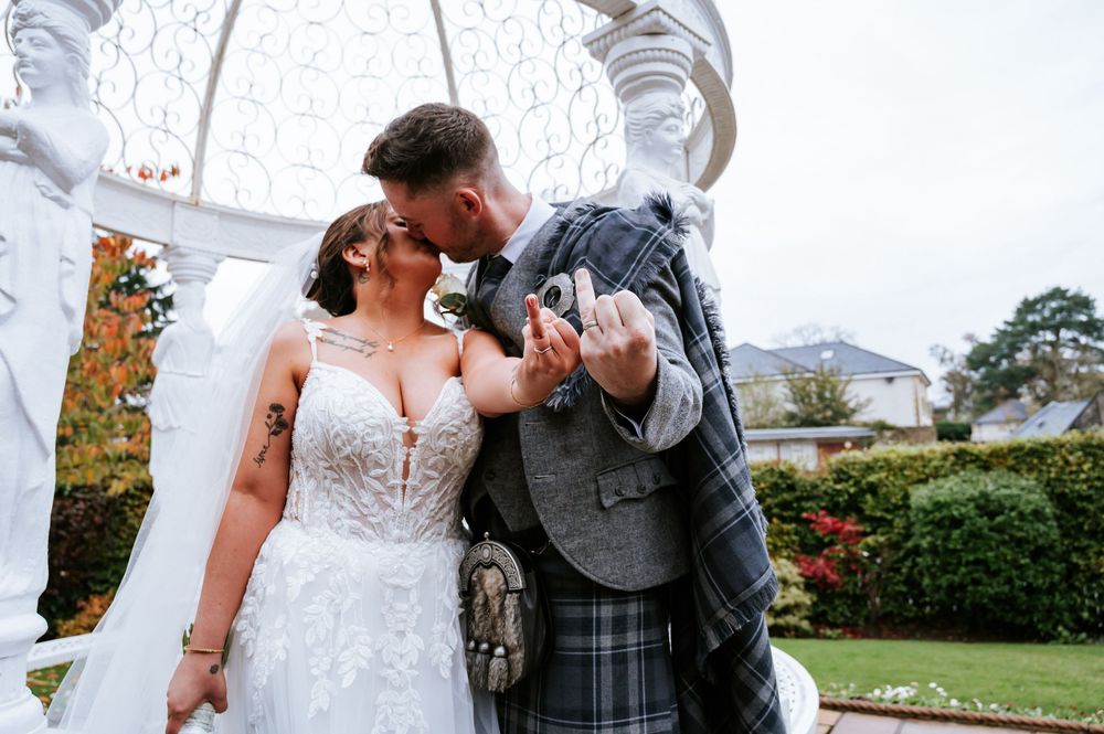 A romantic wedding moment under white pillars with traditional Scottish attire in a garden setting.