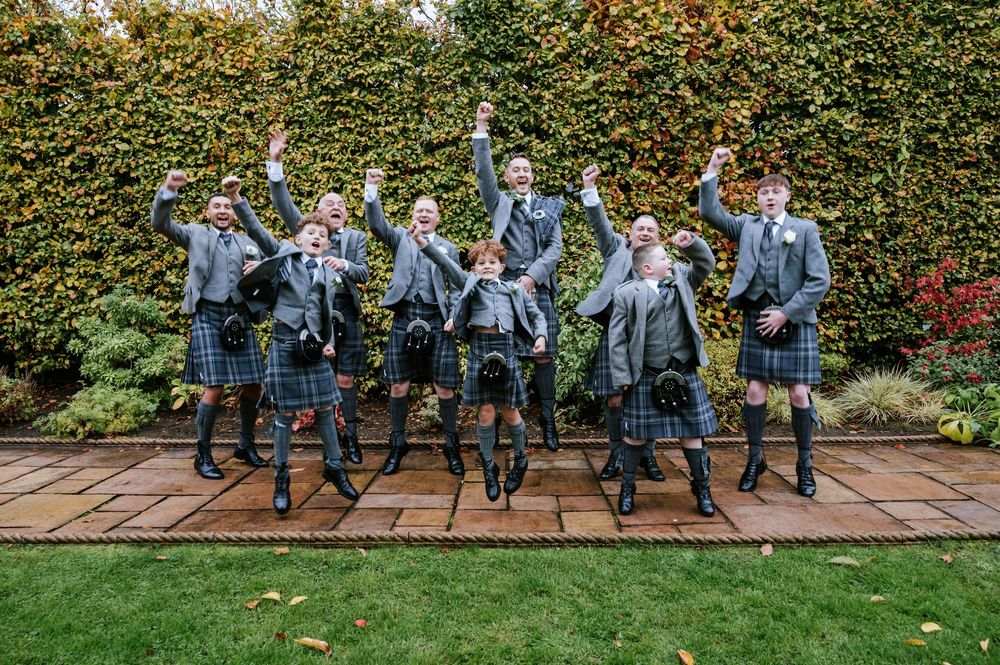 Groomsmen in matching kilts and gray jackets jump in celebration on garden patio.