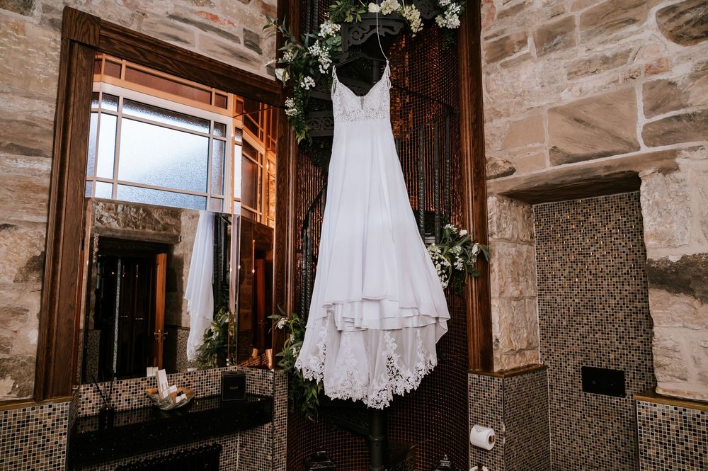A white wedding dress hangs elegantly in a rustic stone-walled room with natural window lighting.