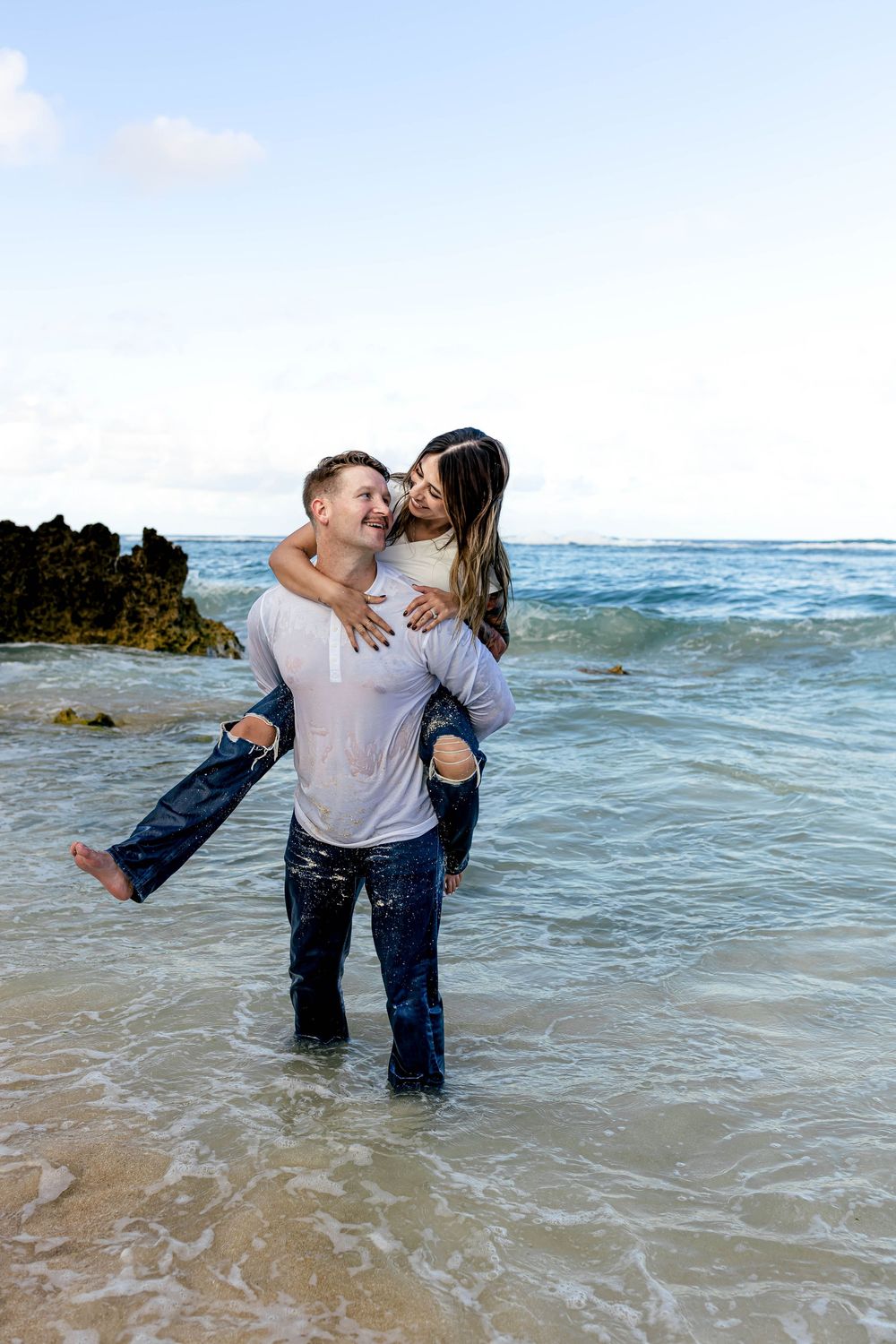Happy couple shares a piggyback ride in ocean waves on a beautiful beach, both laughing and soaking wet.