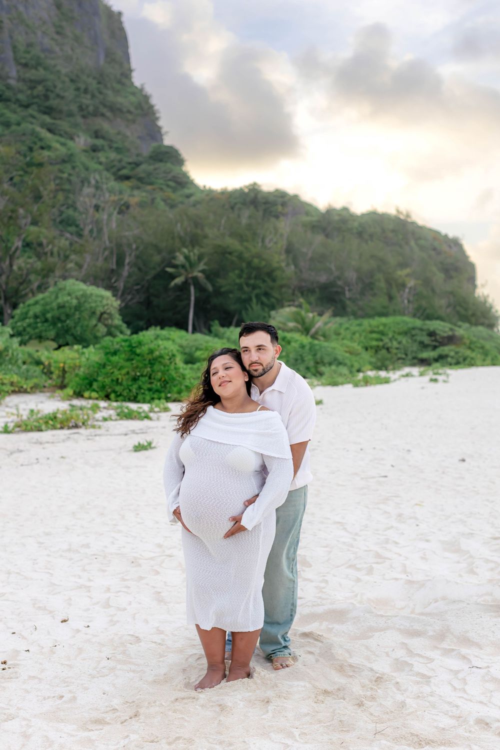 Pregnant couple in white poses on sandy beach with lush green cliffs and dramatic sunset sky behind them.