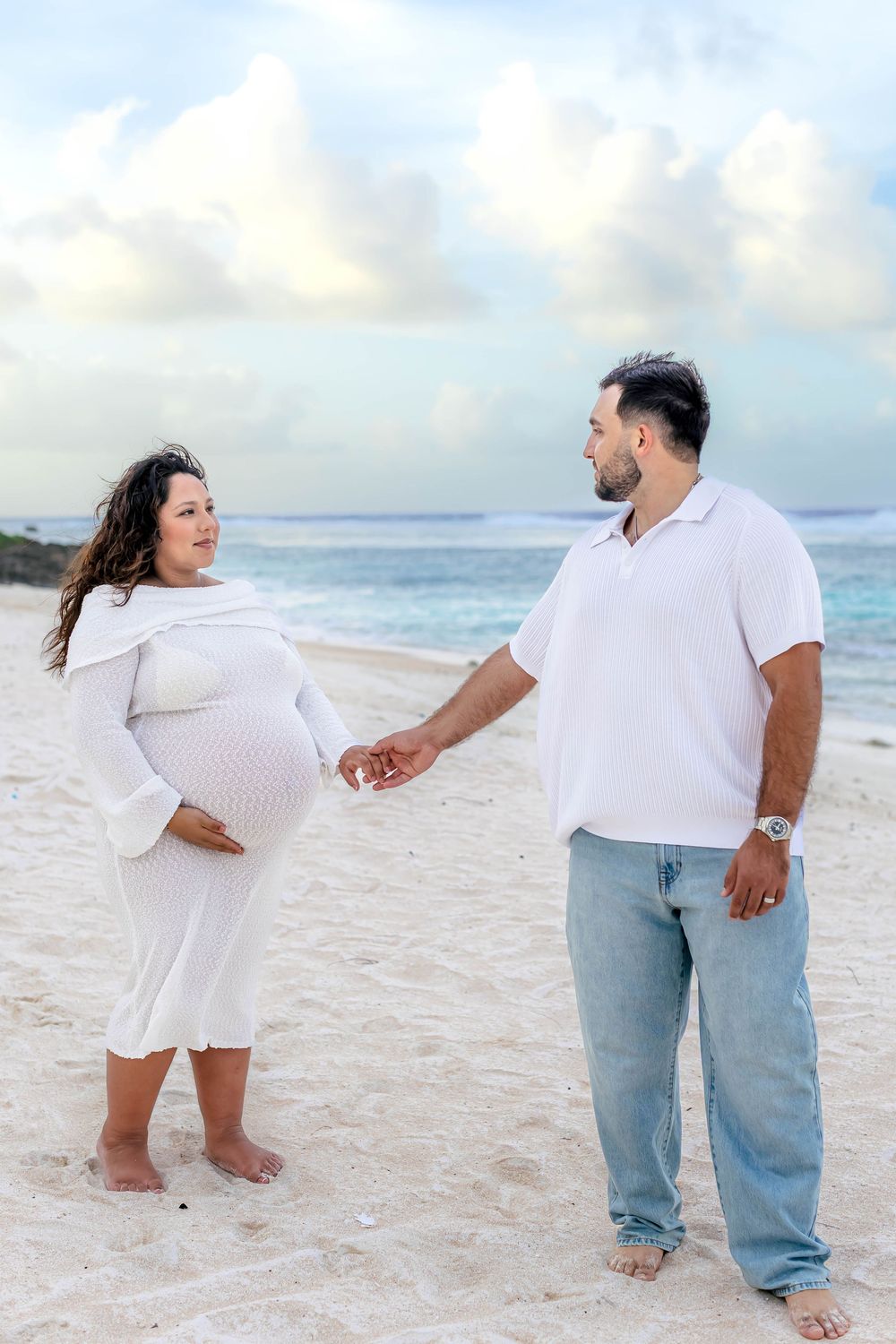 Pregnant woman in white dress holds hands with man on sandy beach, ocean backdrop, maternity photo shoot.