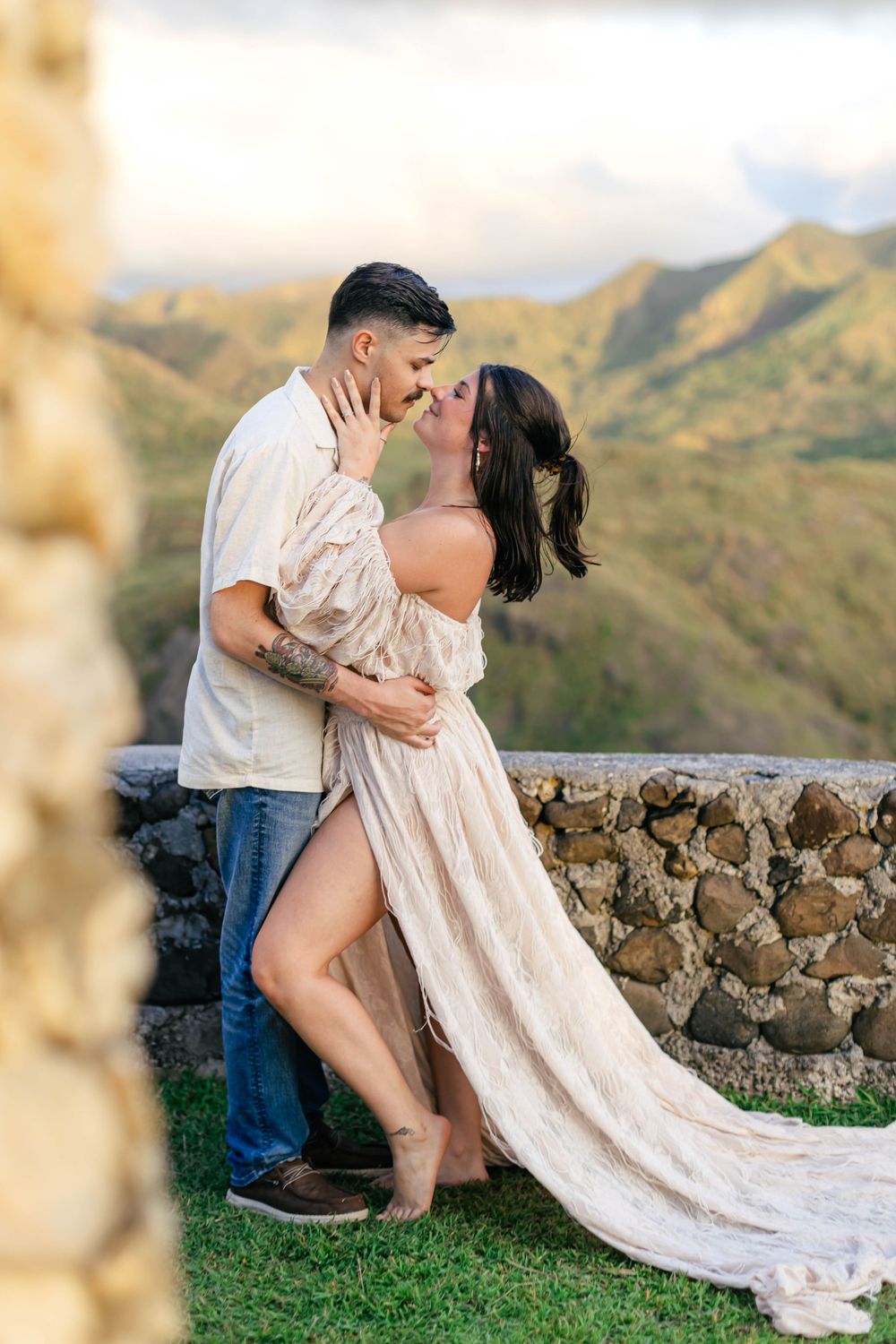 Couple sharing a romantic almost-kiss outdoors, woman in flowing off-shoulder dress, lush green mountains backdrop.