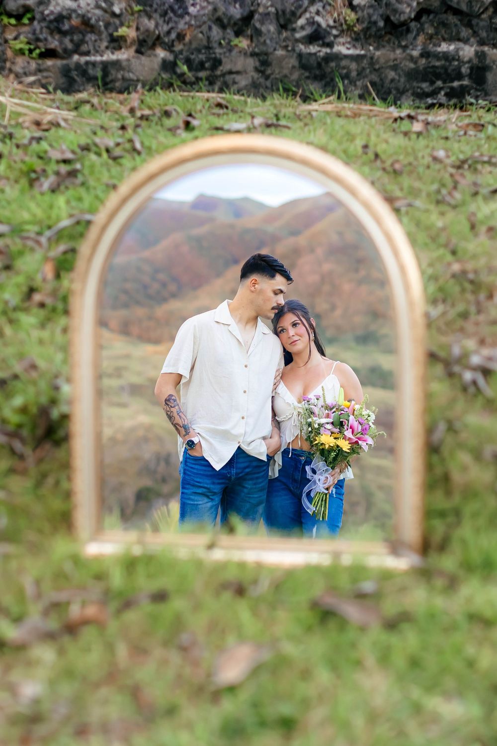 Couple posing in arched mirror reflection with mountain backdrop, woman holding colorful bouquet, man in white shirt.