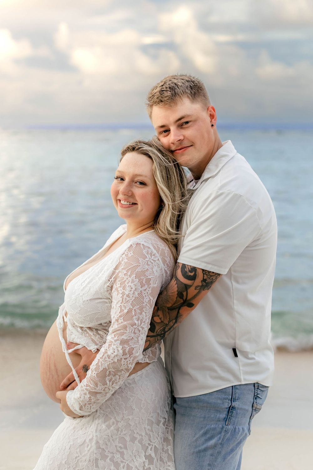 Pregnant couple posing on beach at sunset, woman in white lace maternity dress, man with tattoos in white shirt and jeans.