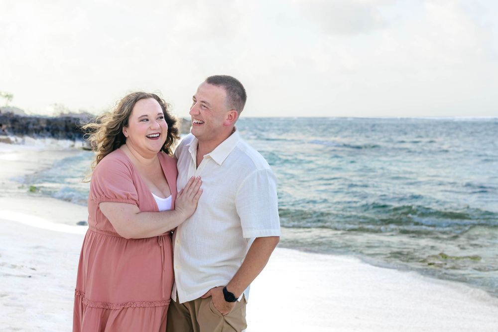 Couple laughing together on a white sandy beach with ocean waves in the background on a sunny day.