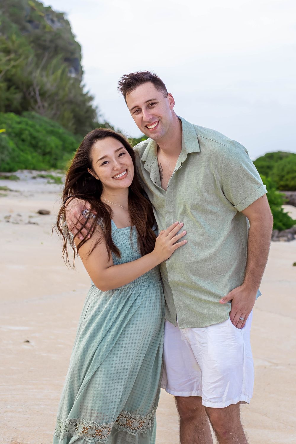 A smiling couple embraces on a sandy beach, woman in teal dress, man in green shirt and white shorts.