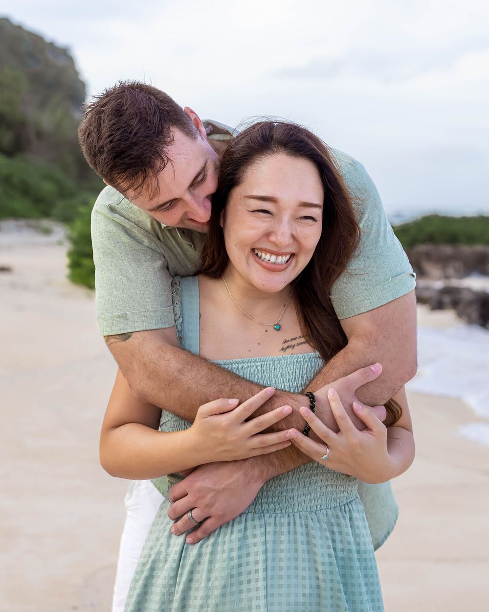 Couple embracing and laughing on a sandy beach, woman in mint dress, man in green shirt, ocean backdrop.