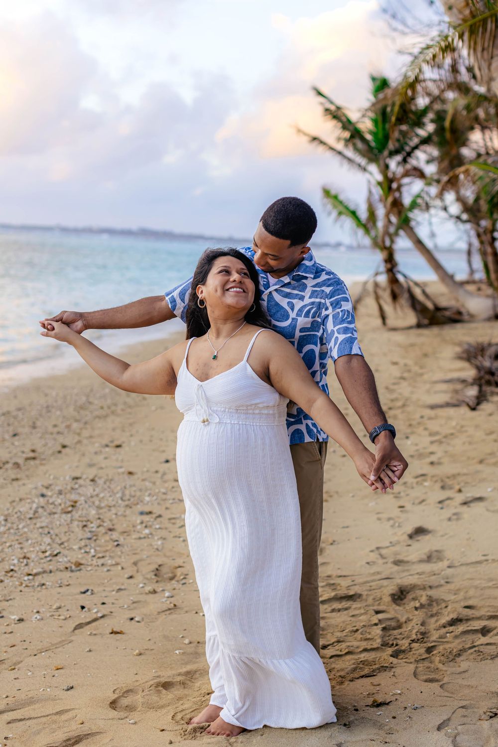 Pregnant woman in white dress dances with man on tropical beach at sunset.