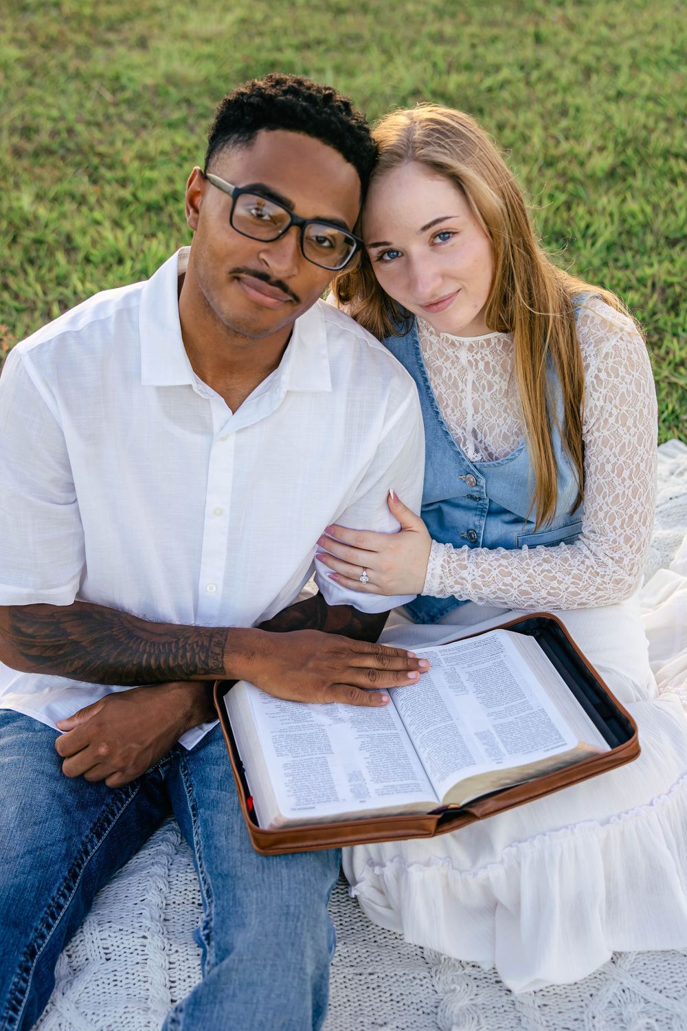 Interracial couple sitting on blanket in grass, sharing a Bible together outdoors.