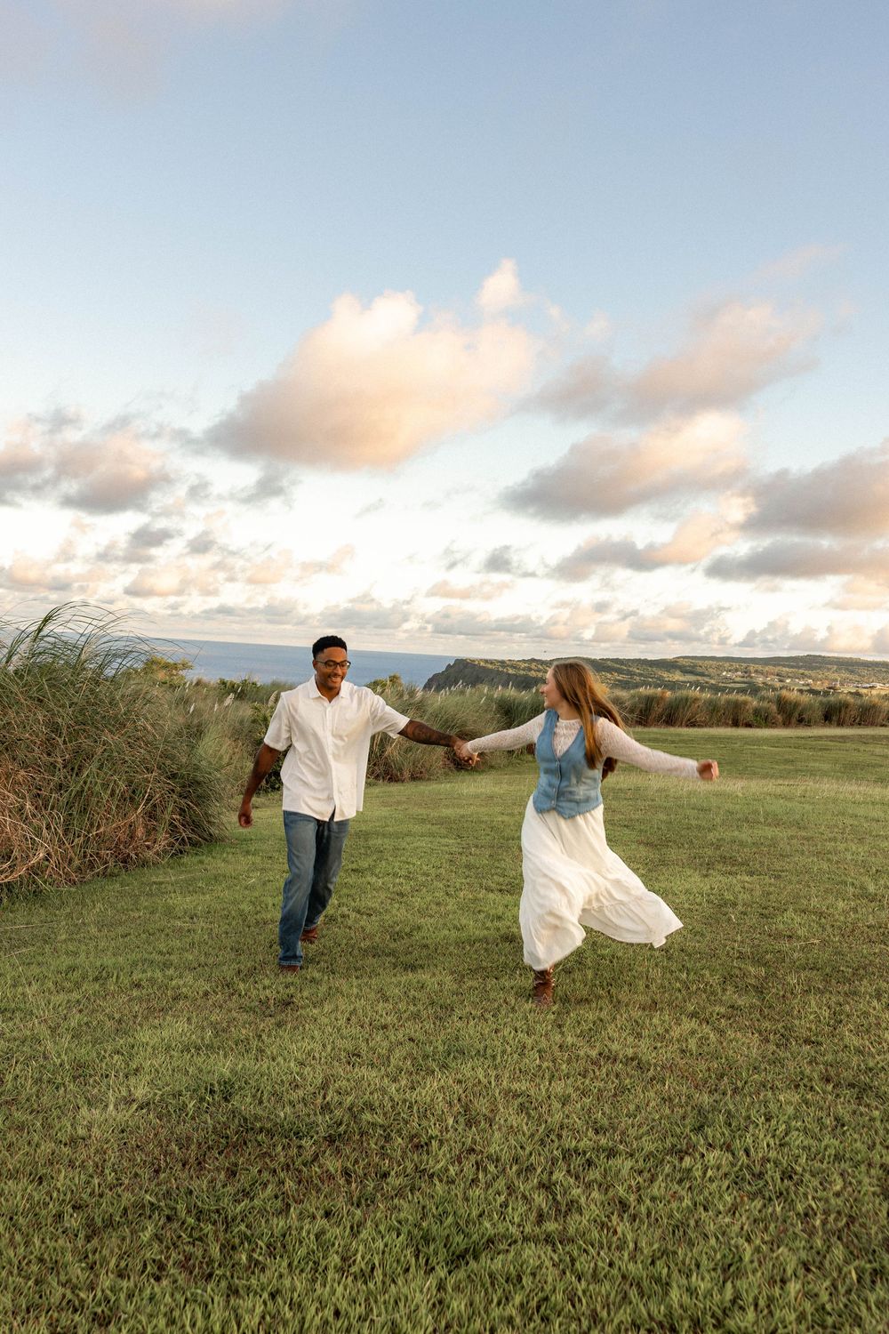 Couple holding hands running through green field at sunset with cloudy sky and ocean view in background.