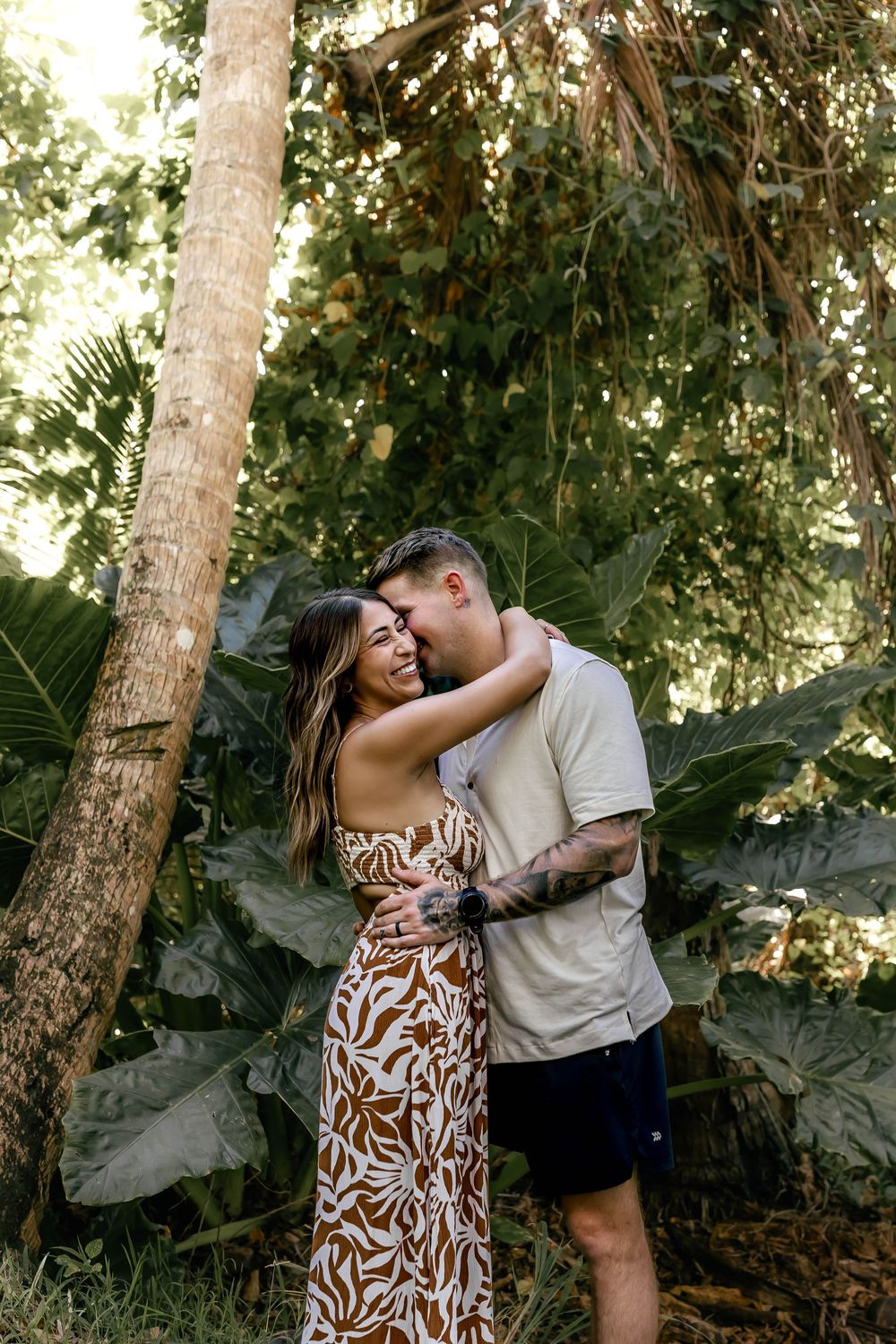 Couple embracing and smiling in a lush tropical garden, woman in floral dress, man in casual attire with tattoos.