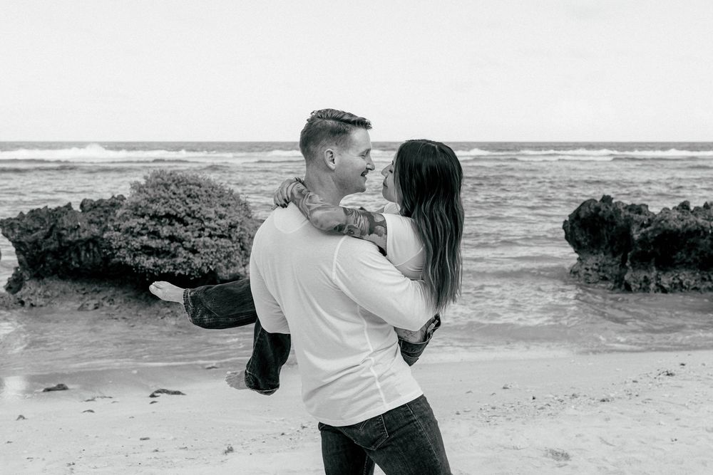 Couple embracing and smiling on rocky beach in black and white romantic photo.