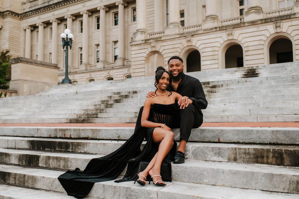 Couple in elegant black attire poses on grand stone steps of historic building during romantic photoshoot.