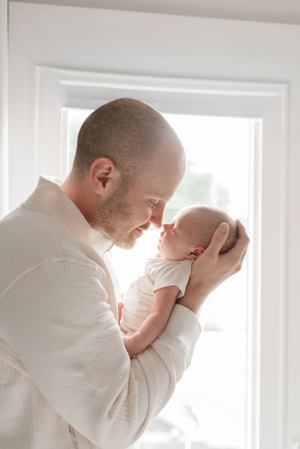 Father and baby by a bright window as their faces touch, in a sweet moment capture by Jess GC Photography, Toronto newborn photographer