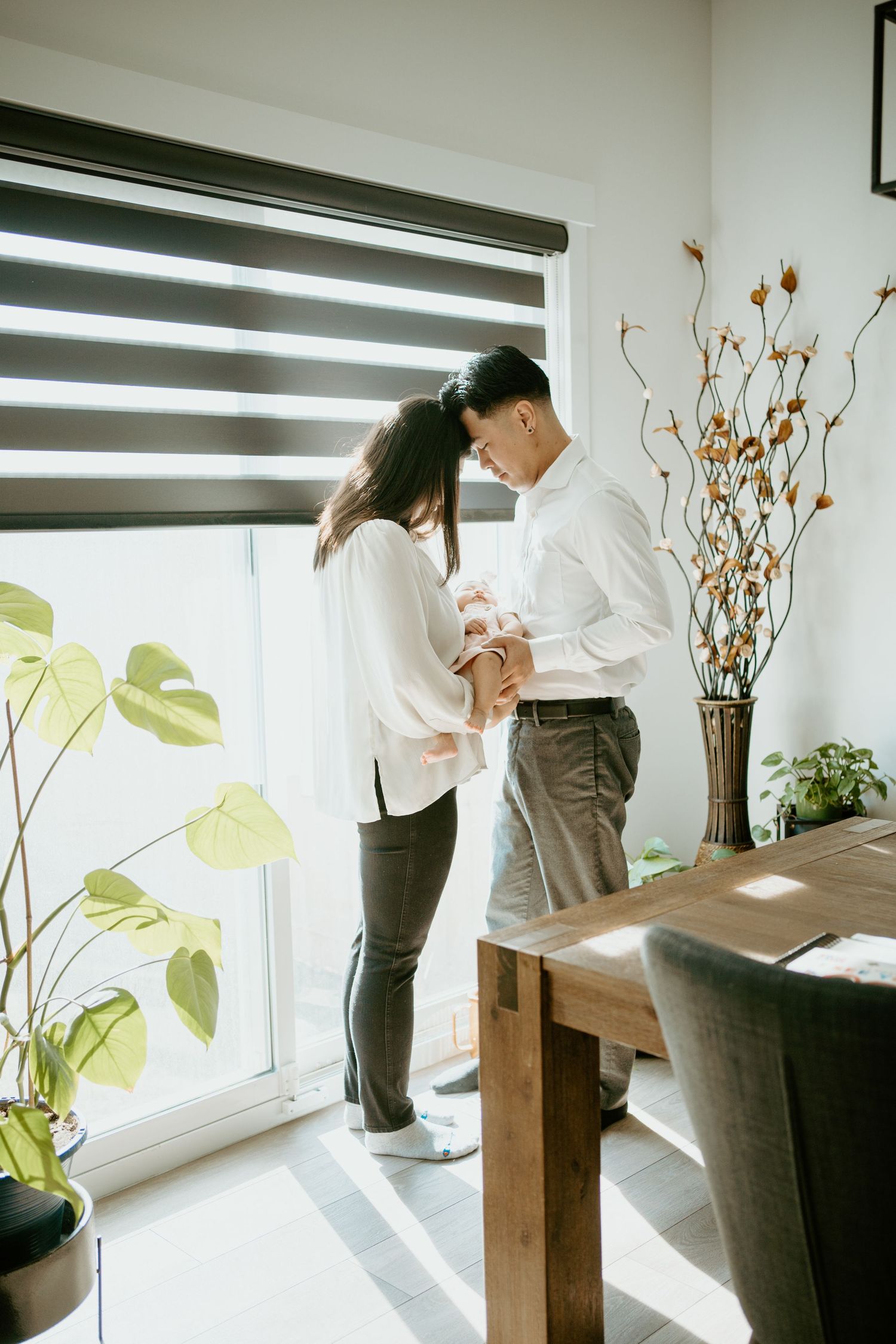 A couple shares an intimate moment near a window with houseplants and modern zebra blinds in a bright interior setting.