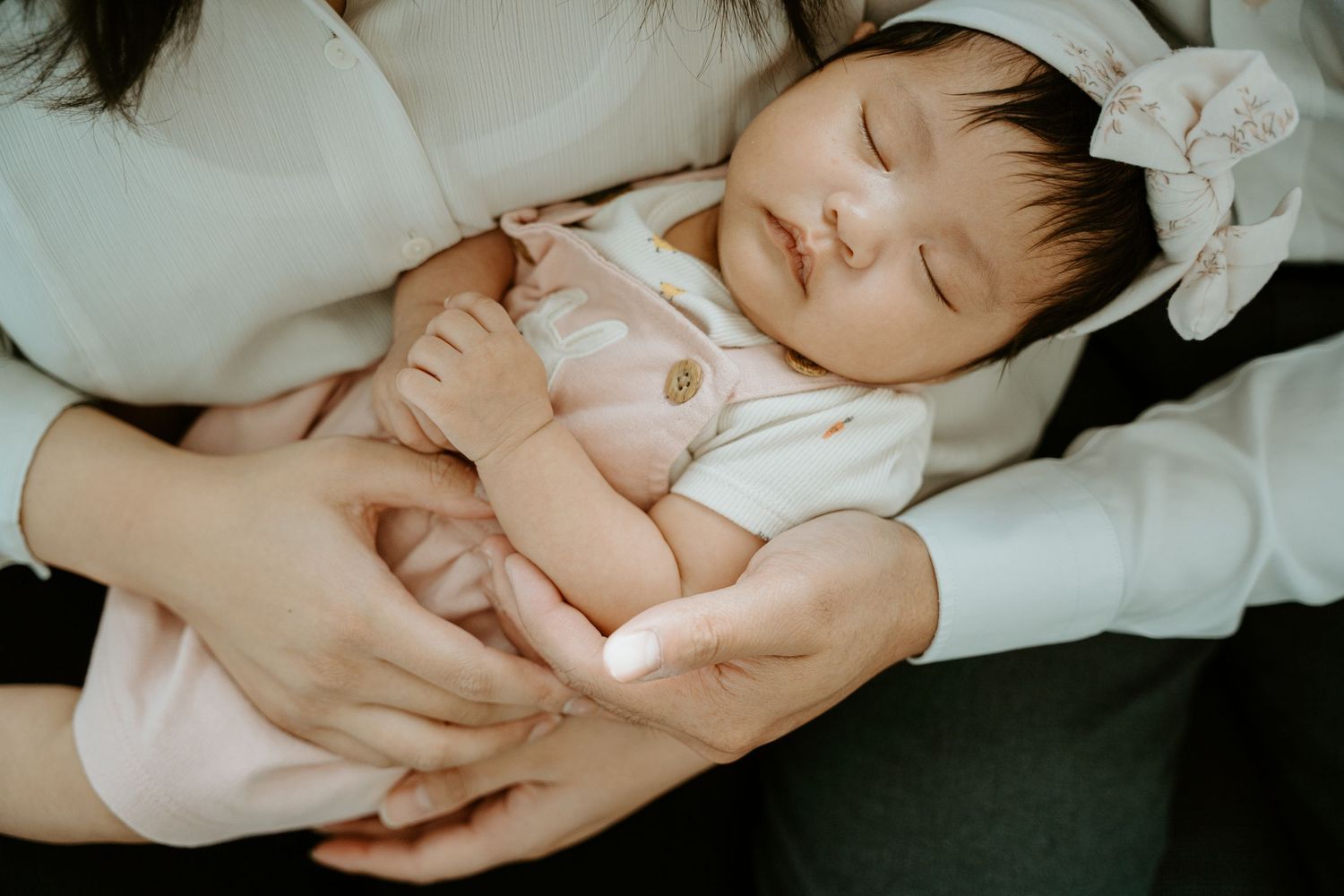 A sleeping baby in a white outfit is cradled gently in arms while resting peacefully.