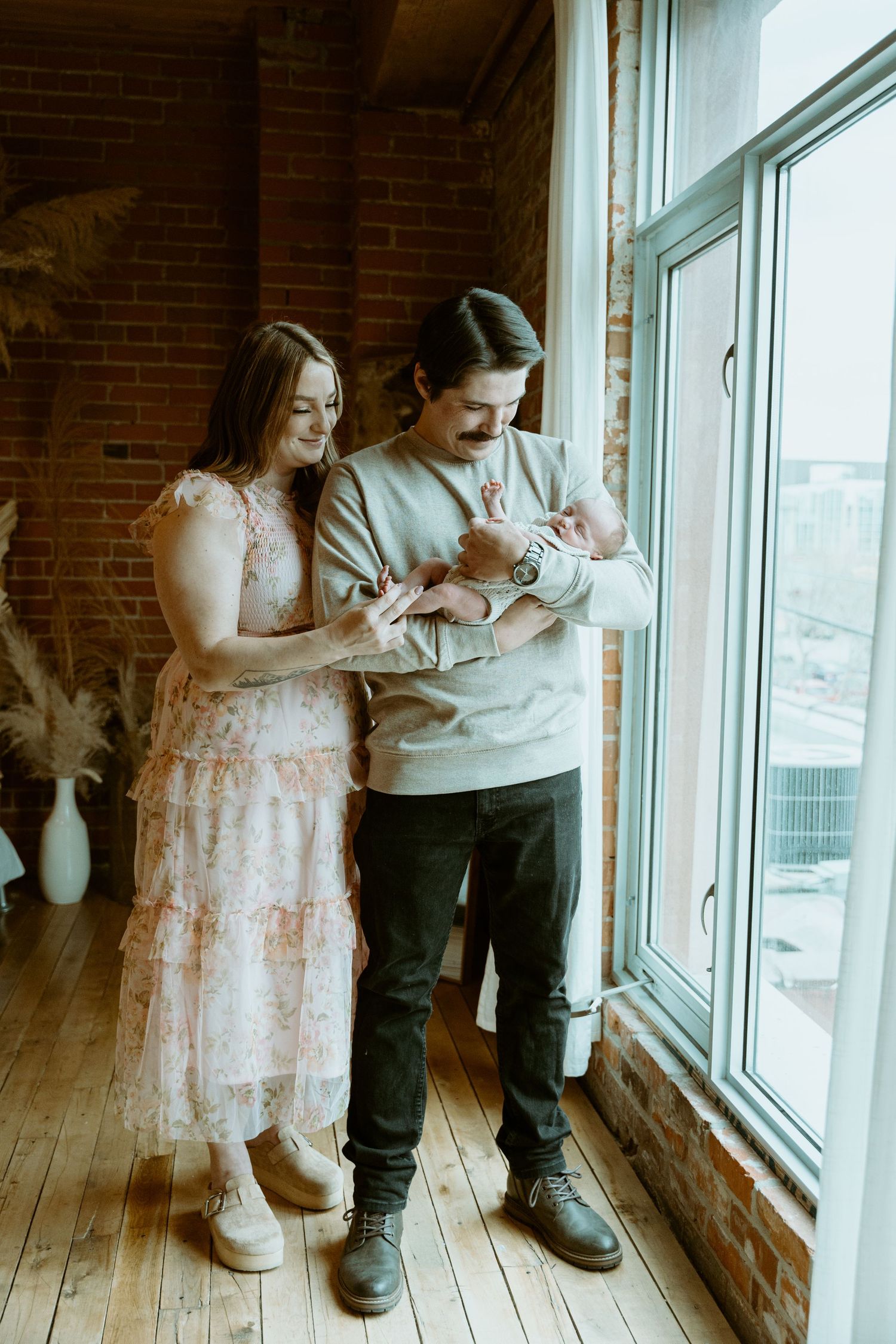 Two people in casual attire sit together by a bright window in a room with exposed brick walls and hardwood floors.