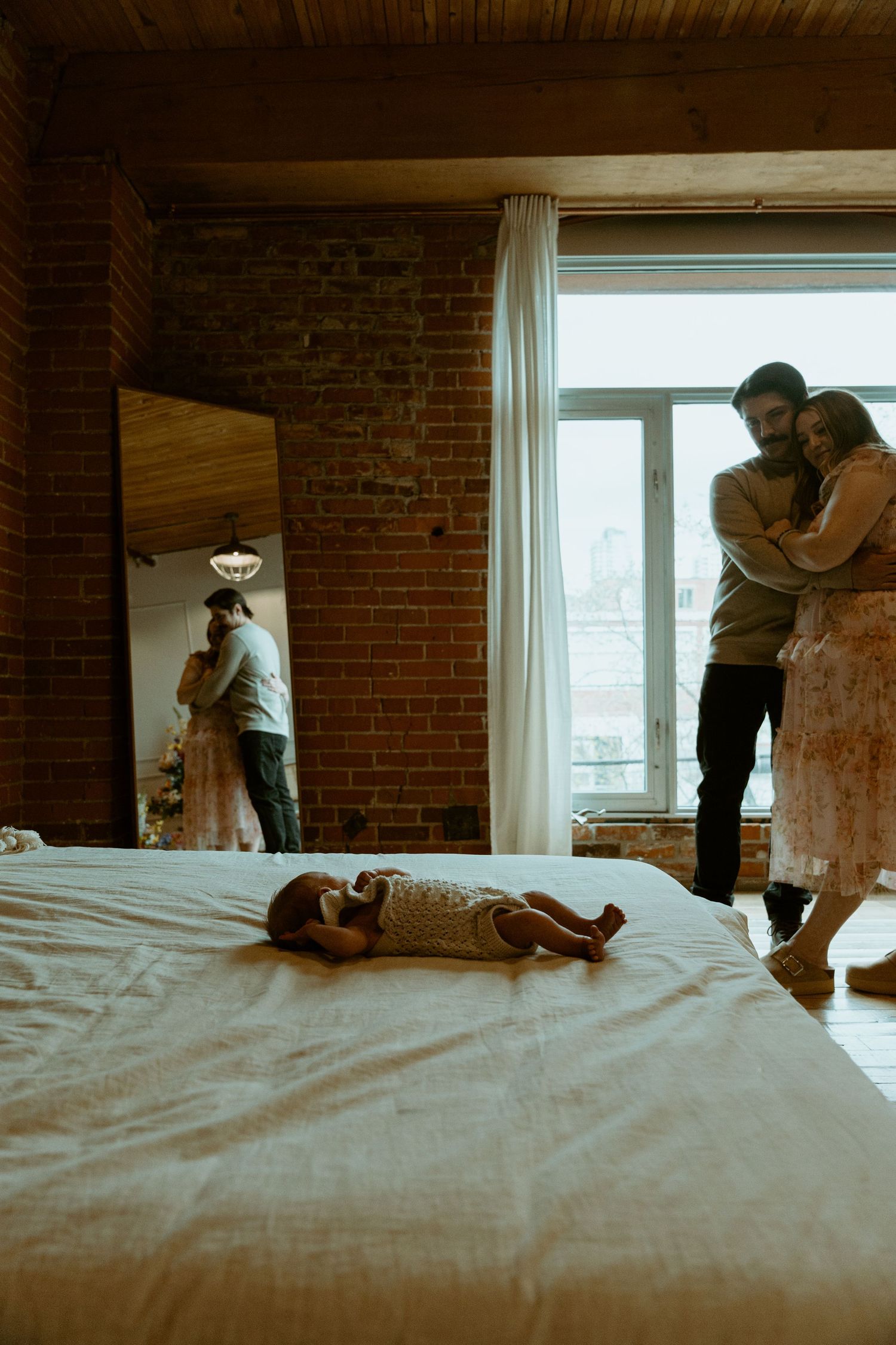 A baby lies on a bed in a rustic brick-walled room while others stand near a bright window.