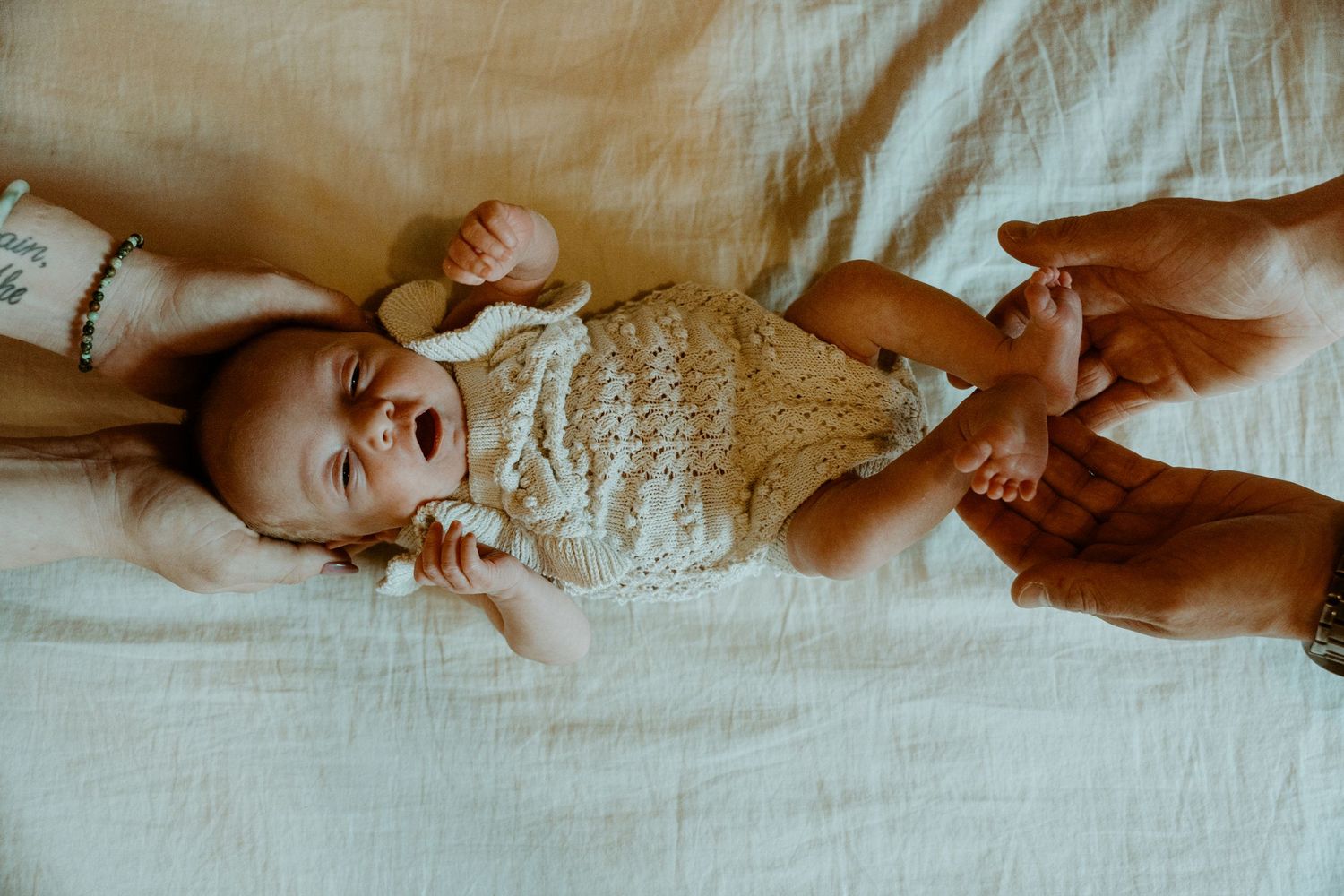 A baby wearing a cream knit romper lies on white bedding while hands gently care for them.