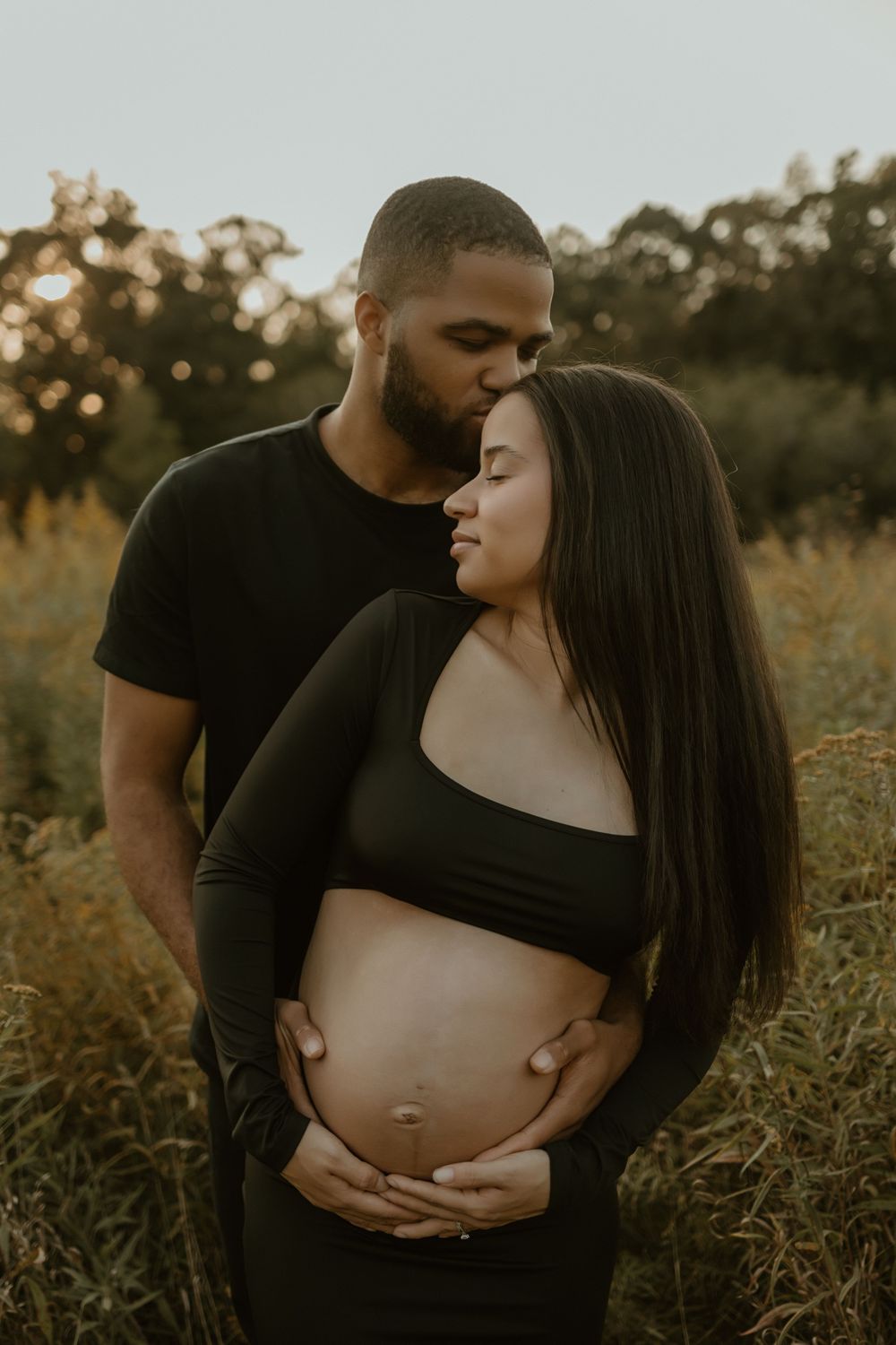 Romantic maternity photos captured in warm evening light with couple dressed in black embracing.