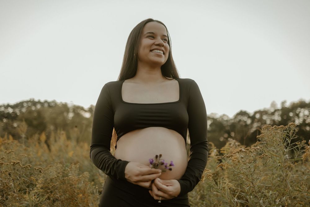 A woman in a black top poses outdoors against a light background during a maternity photoshoot.