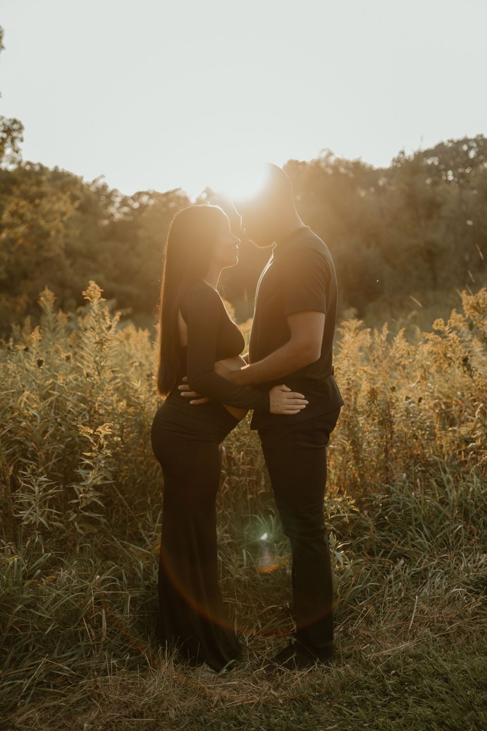 A silhouetted couple embraces in a sunlit meadow during golden hour as the sun sets behind them.