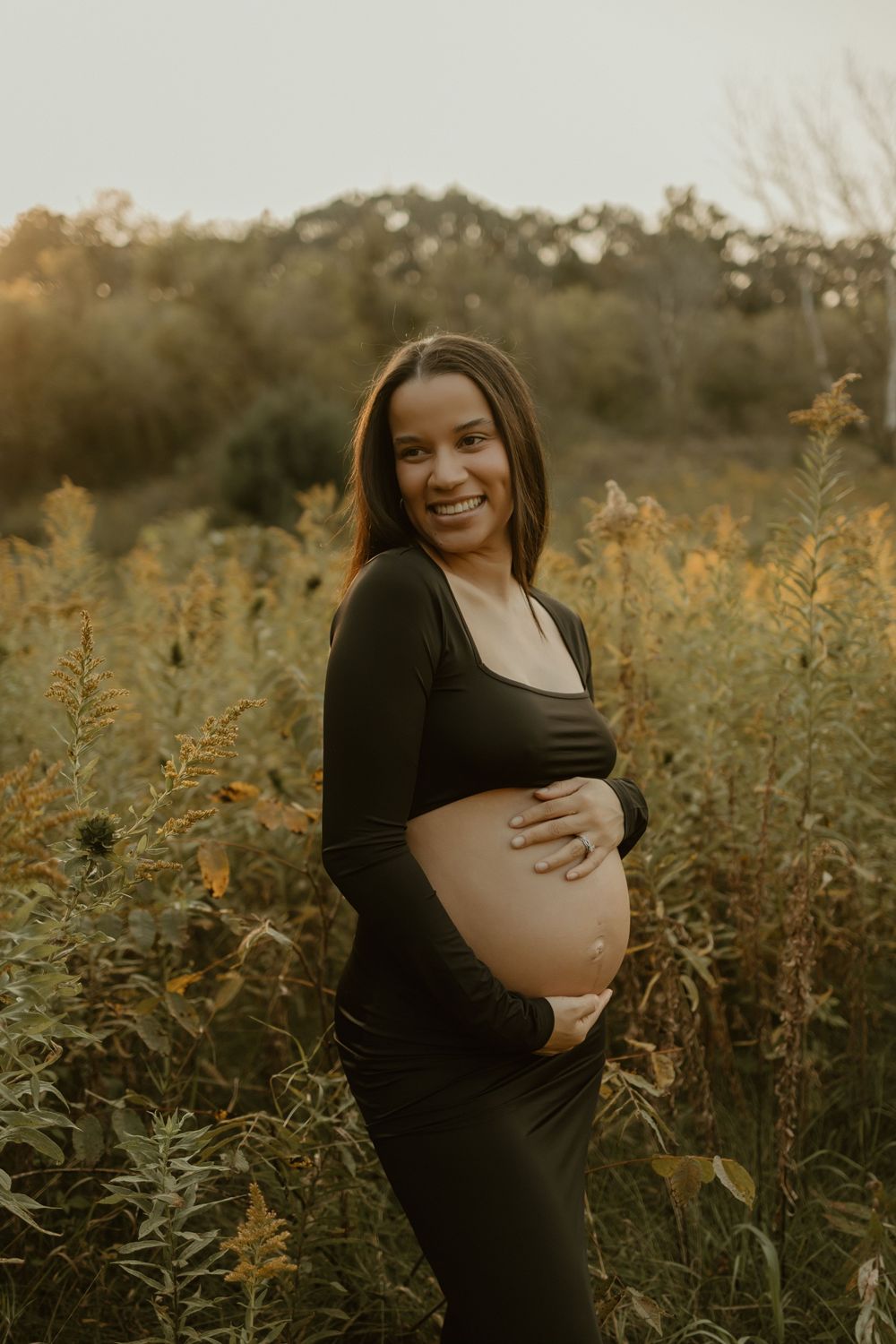 A maternity photoshoot in a golden field at sunset shows a woman in black caressing her pregnant belly.