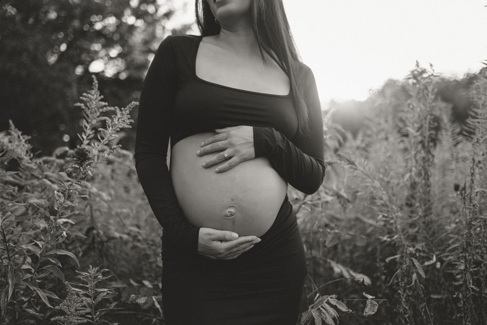 Black and white photo of an expectant mother in a dark dress cradling her baby bump in a natural outdoor setting.