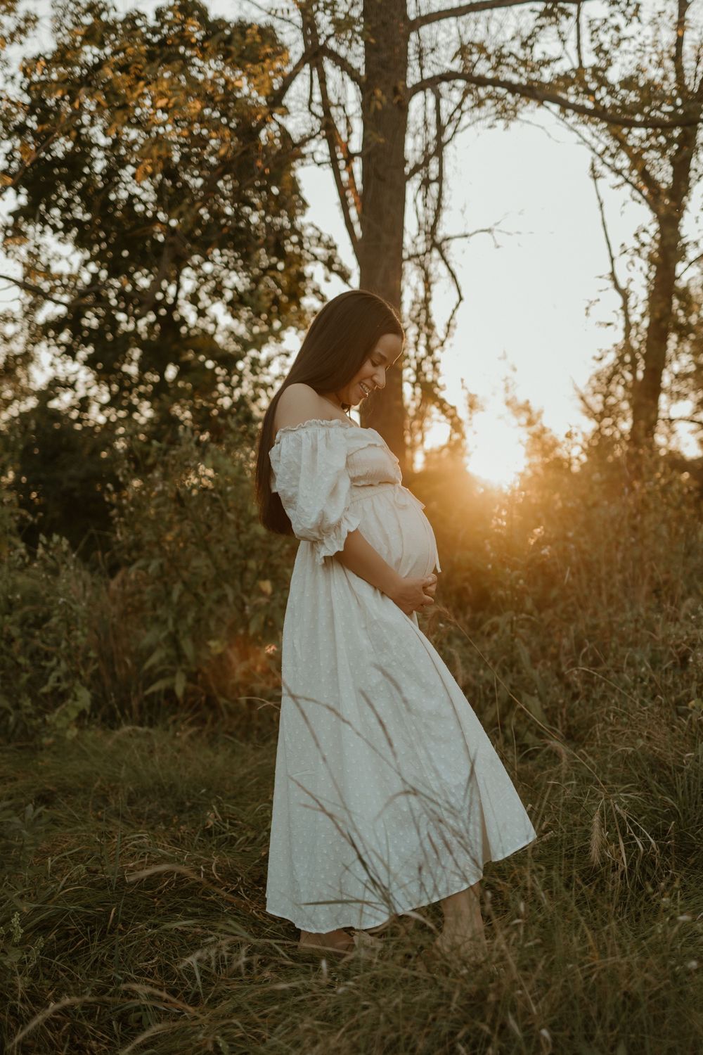 A person in a flowing white dress walks through tall grass at sunset in a wooded area.