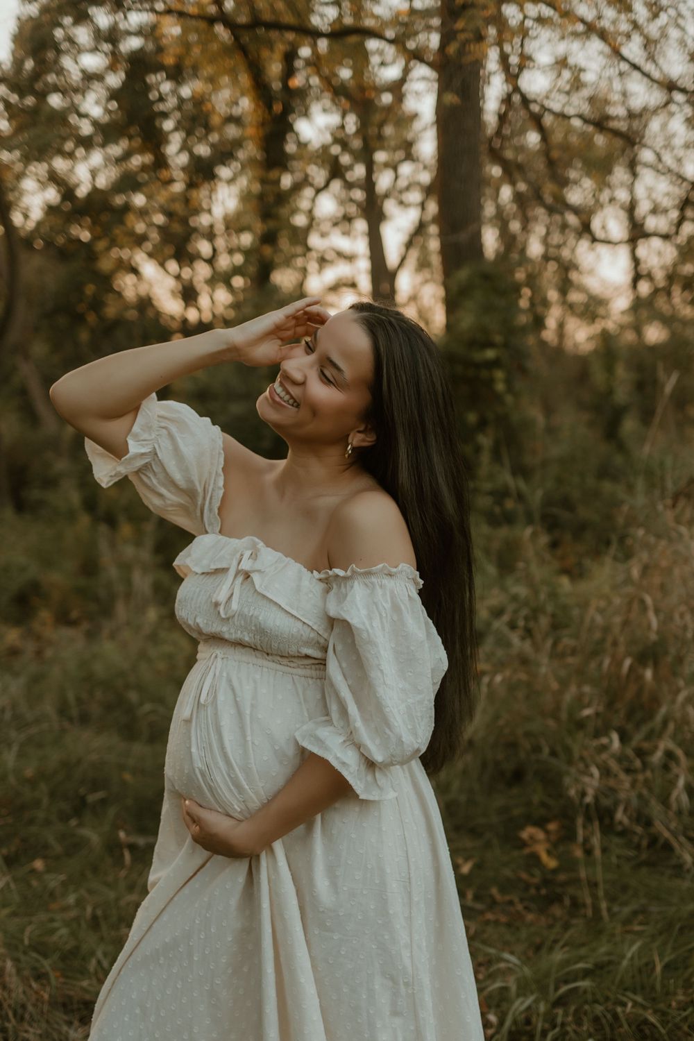 Two women in white dresses embrace and laugh together in a sunlit forest setting during golden hour.